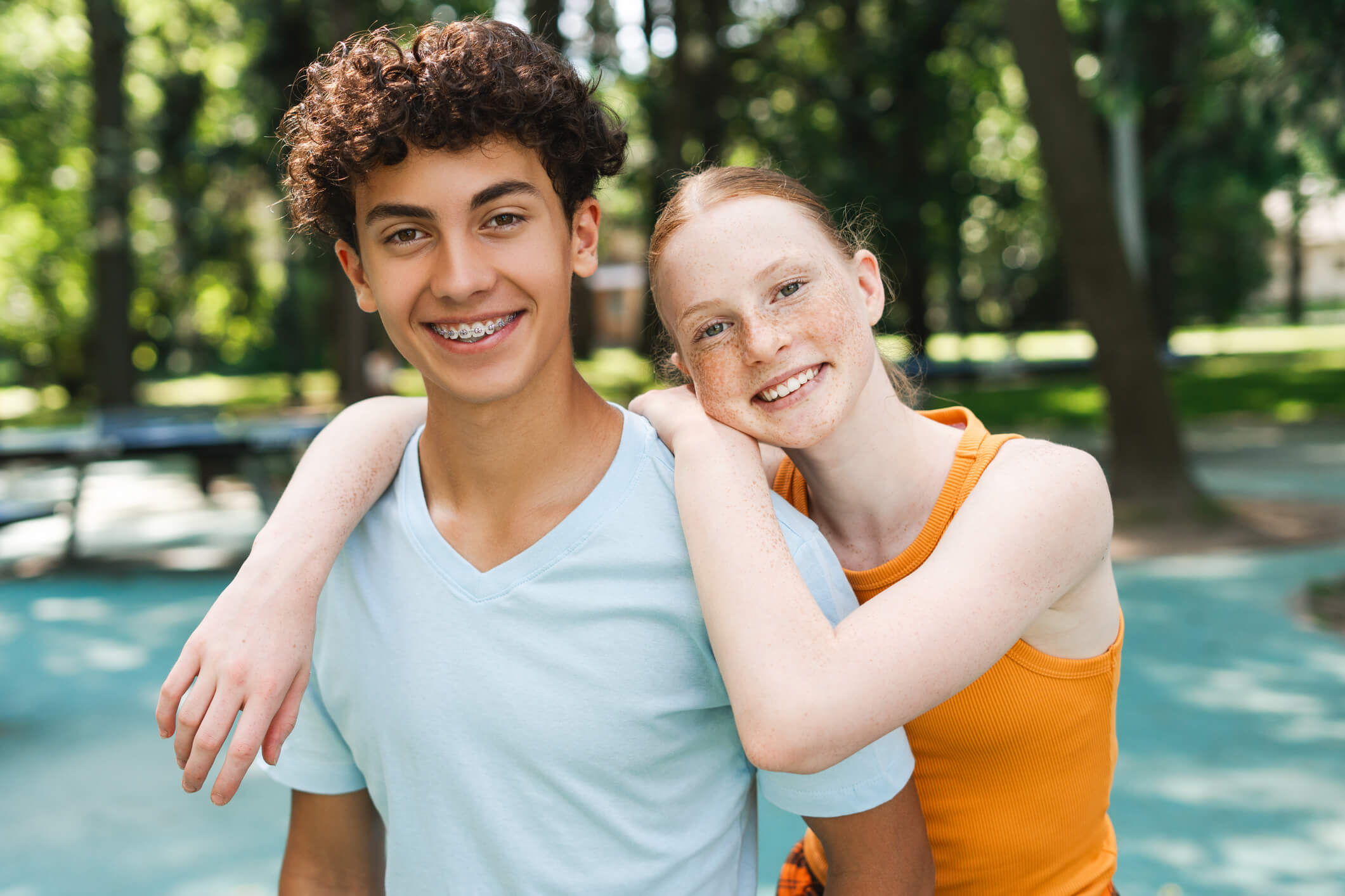 Smiling teen boy with braces and a freckled girl resting her chin on his shoulder in a park