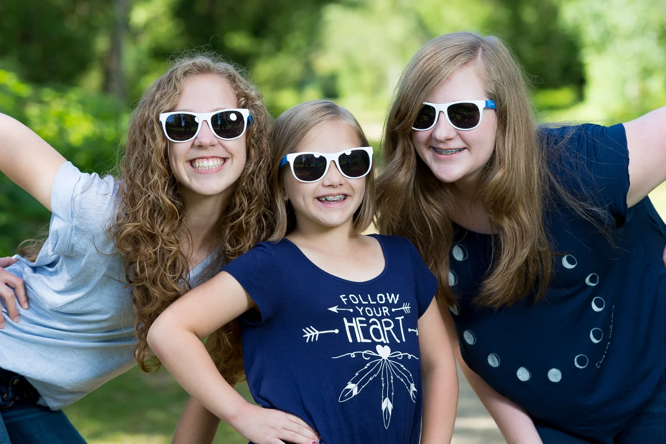 Three smiling girls wearing white sunglasses pose together outdoors