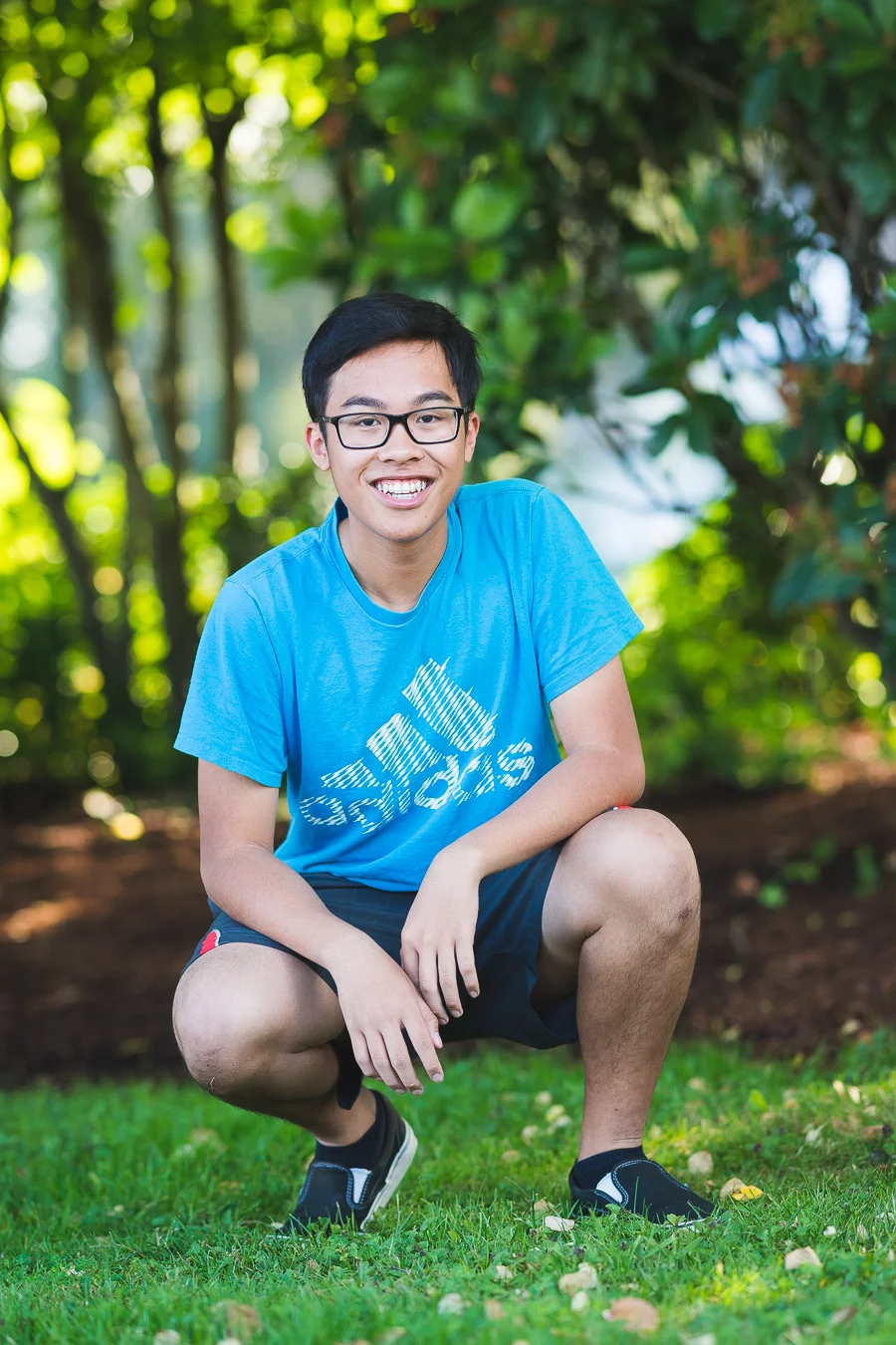 Smiling teen in glasses and blue t-shirt crouching on grass outdoors