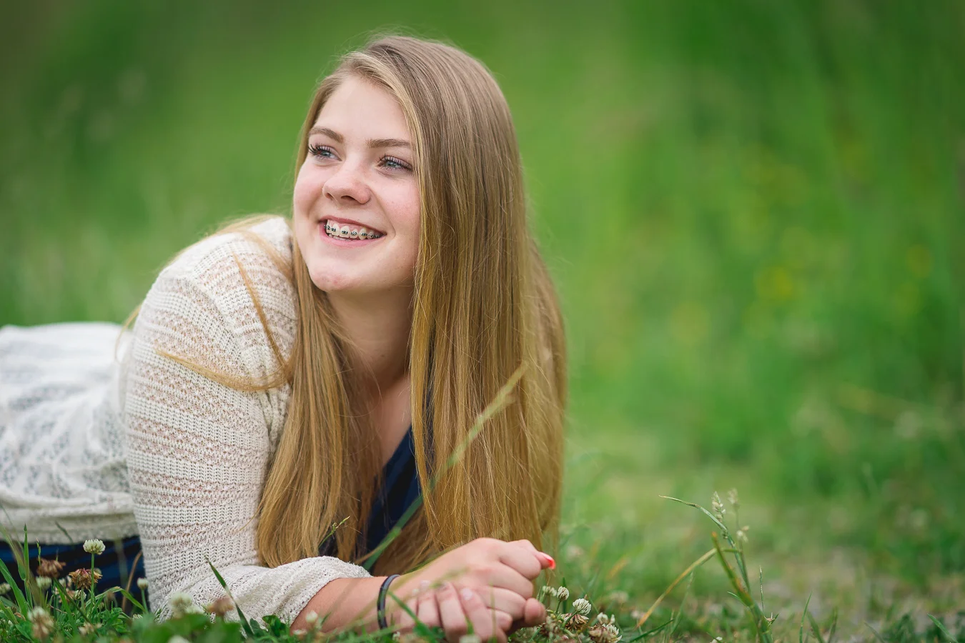 Smiling teen girl with braces lying in grass, long blonde hair and cream sweater