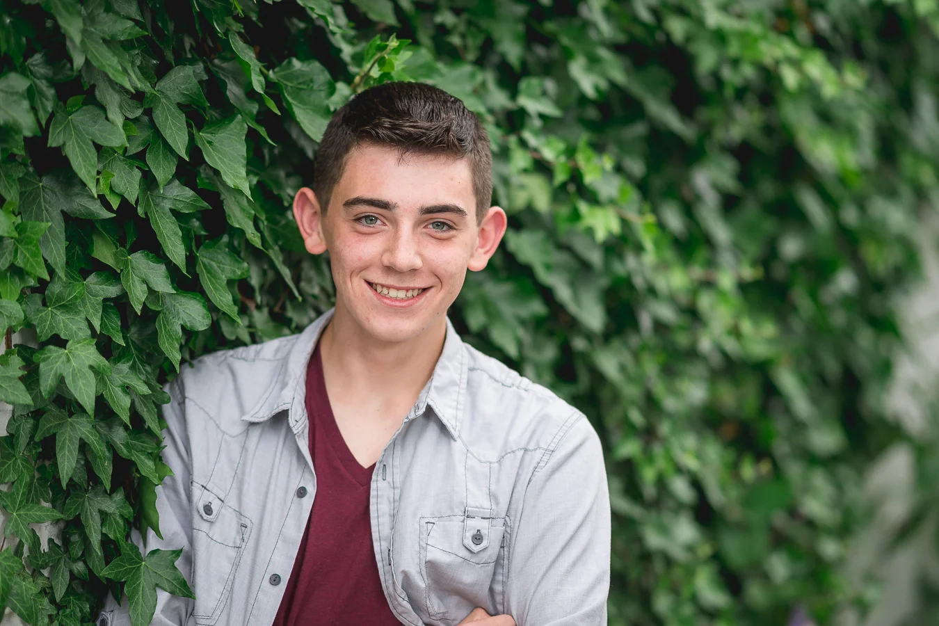 Smiling teen boy in denim shirt standing against a green ivy wall