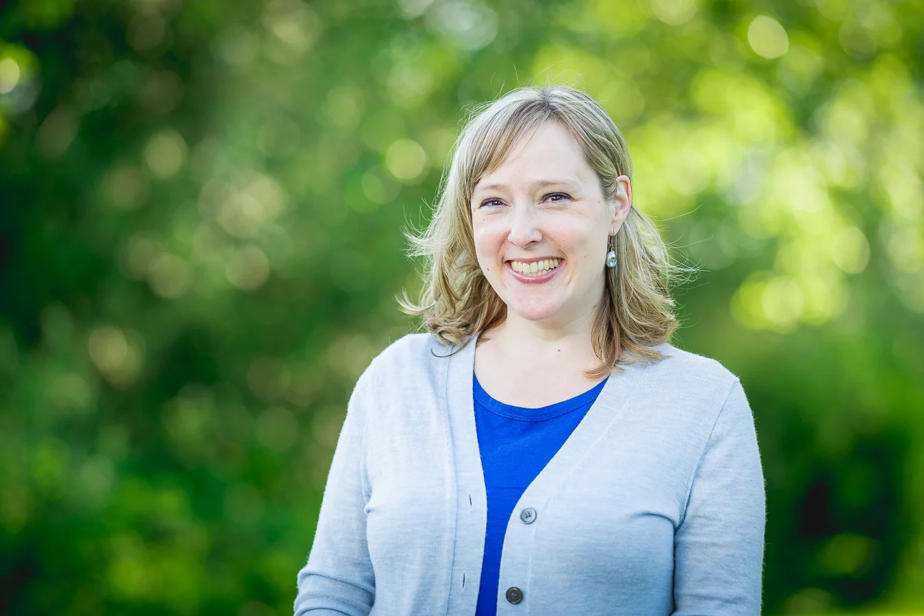 Smiling woman with blonde hair wearing a gray cardigan over a blue top, outdoors with greenery