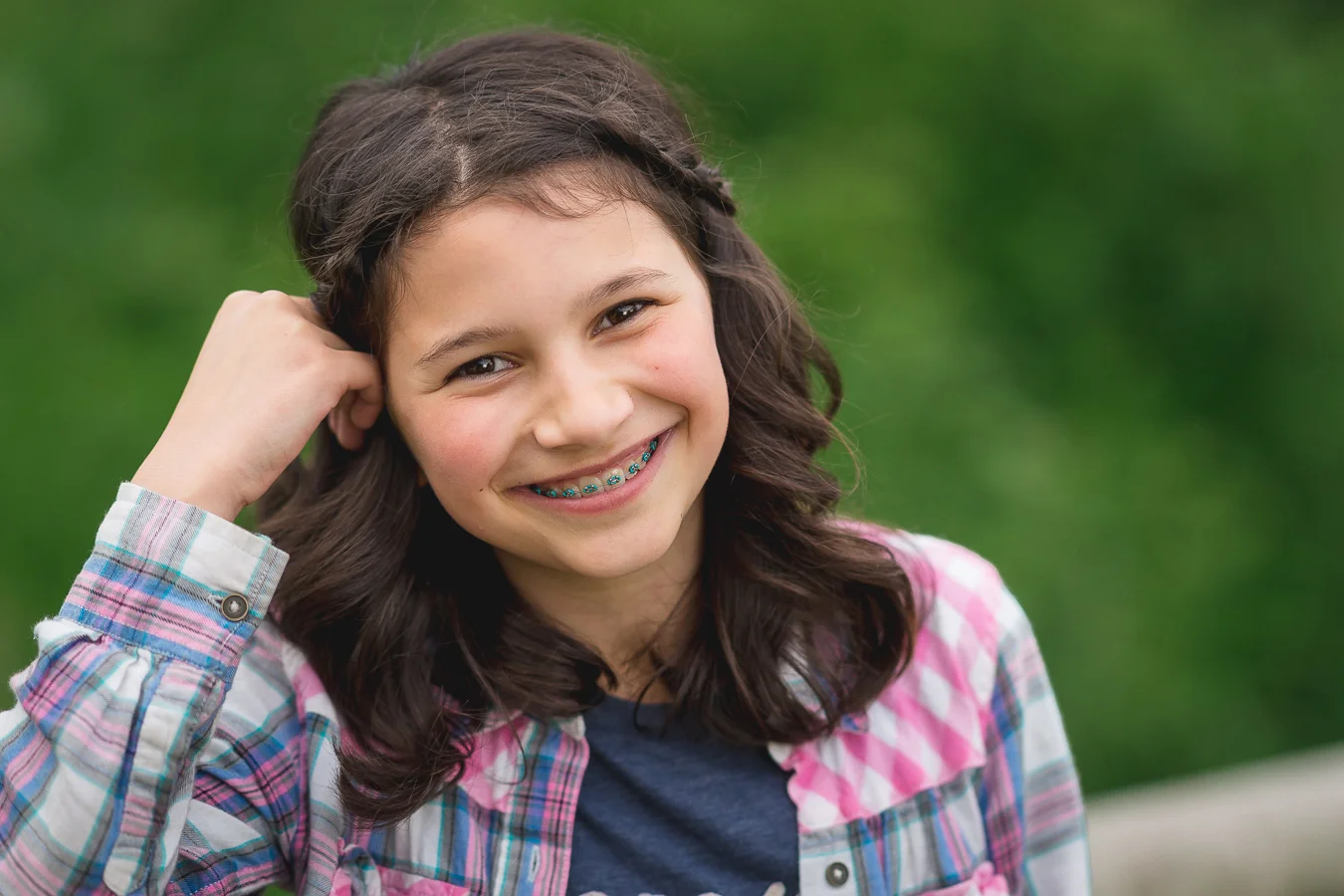 Smiling girl with braces and wavy brown hair wearing a pink and blue plaid shirt outdoors