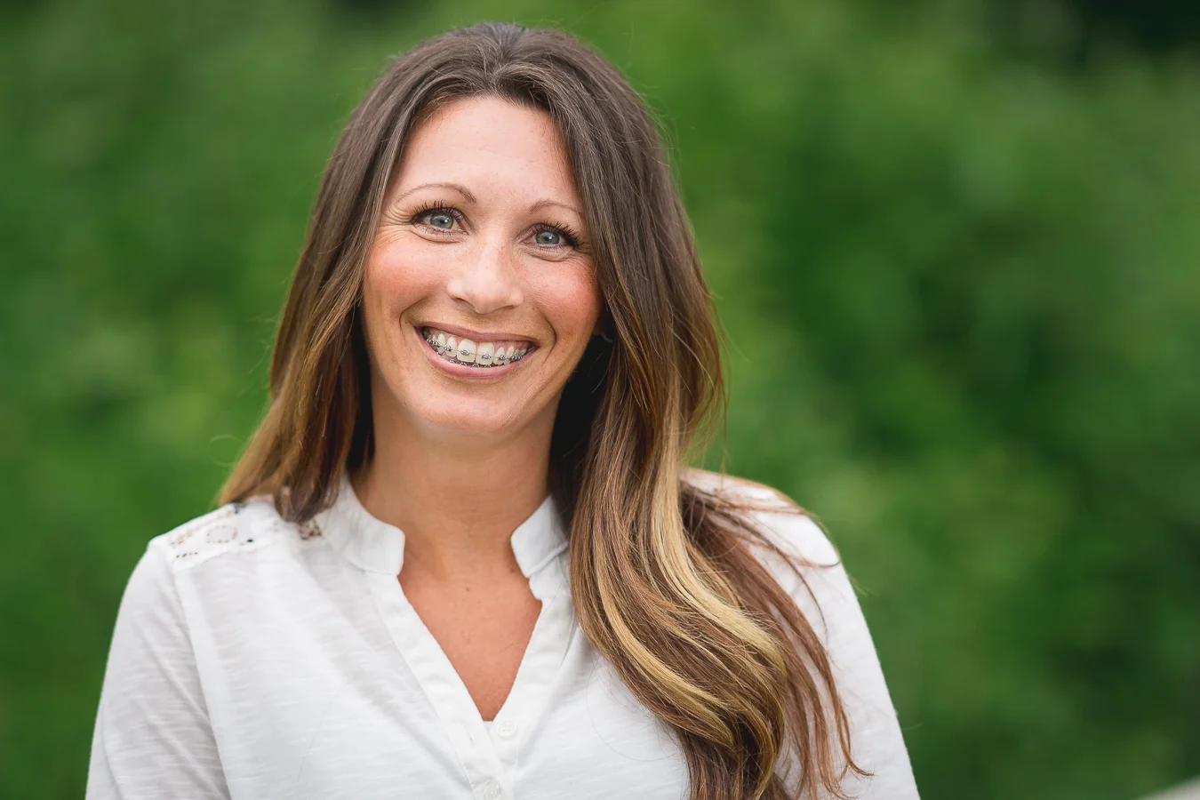 Smiling woman with metal braces and long brown hair, wearing a white blouse outdoors