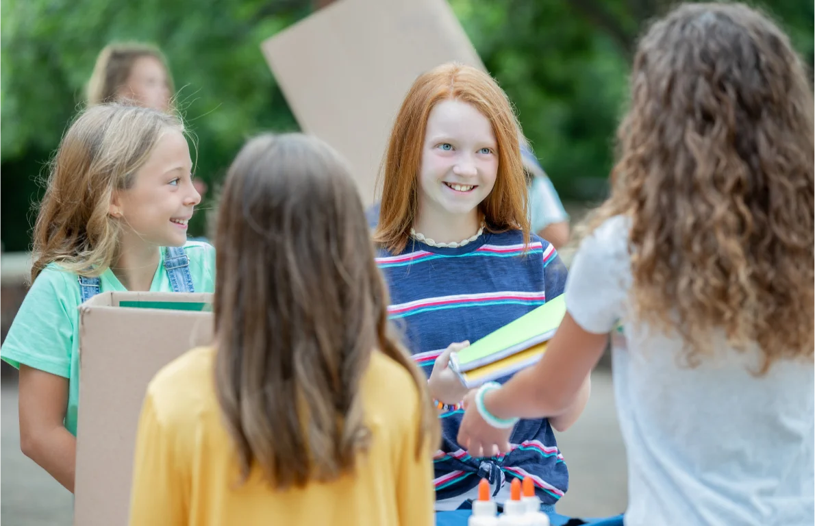 Smiling kids gathered outdoors with cardboard boxes, notebooks, and glue bottles for a donation drive