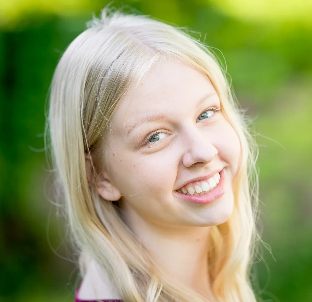 Smiling blonde teenage girl with straight white teeth outdoors against a green background