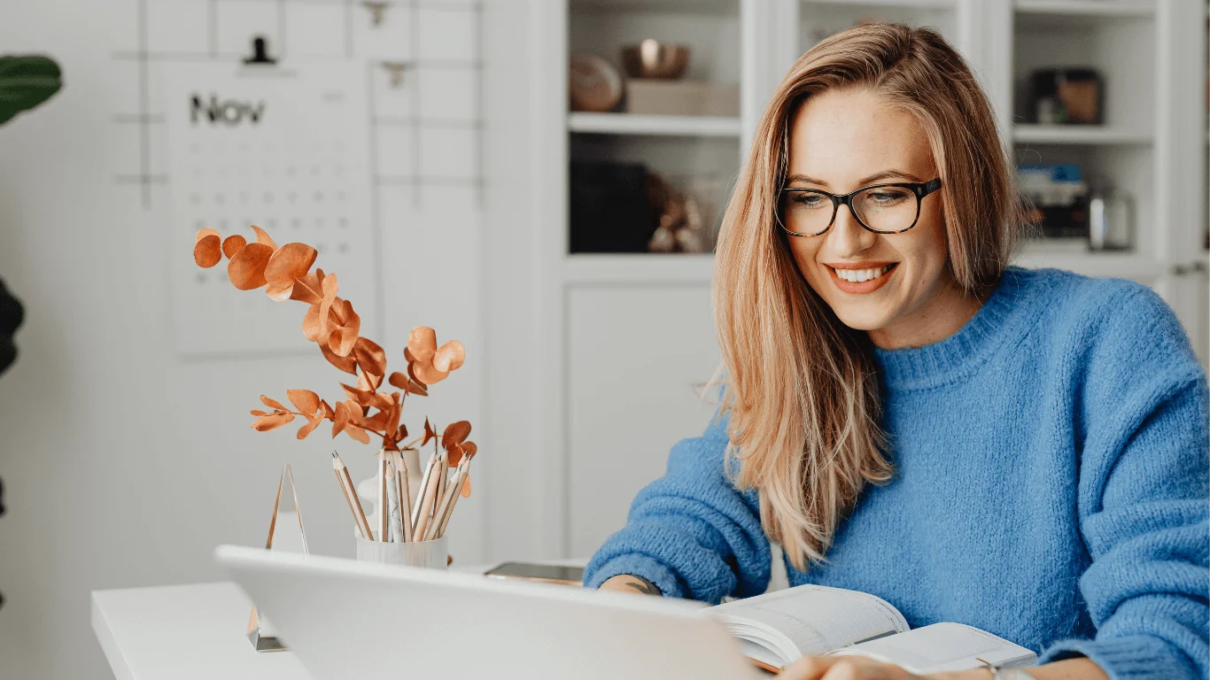 Smiling woman in glasses and blue sweater reading a book at a desk