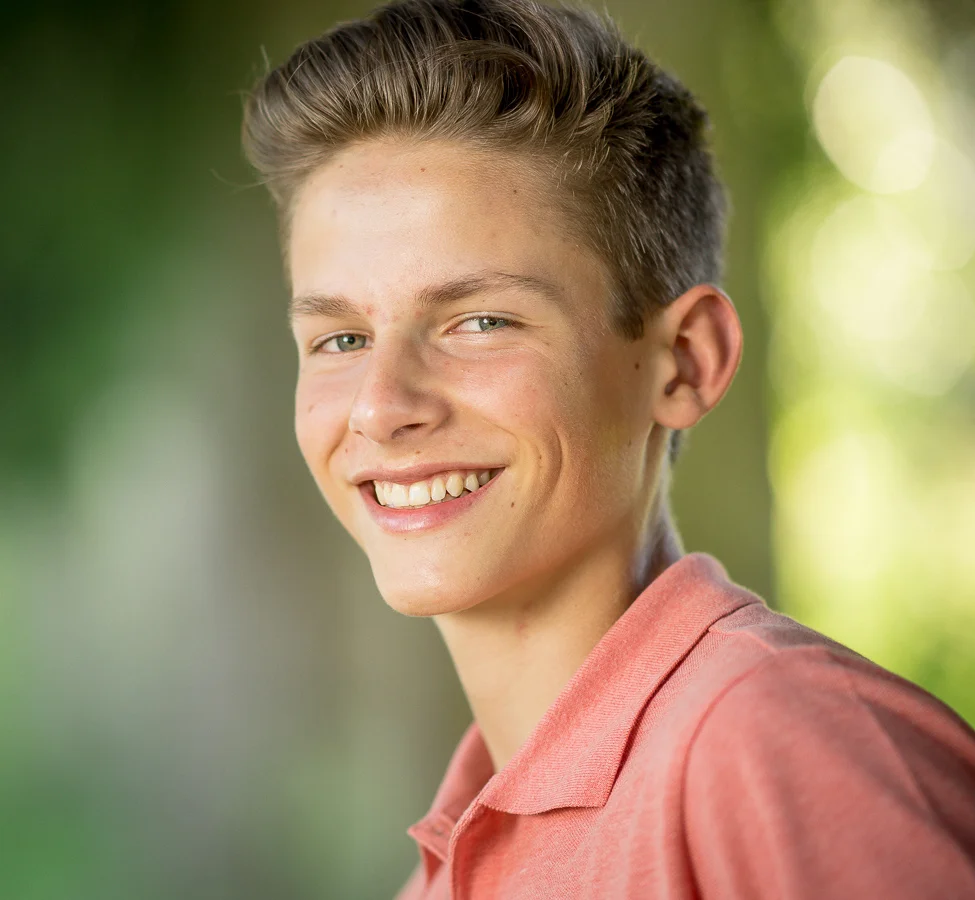 Smiling teenage boy in a coral polo shirt outdoors with a green leafy background