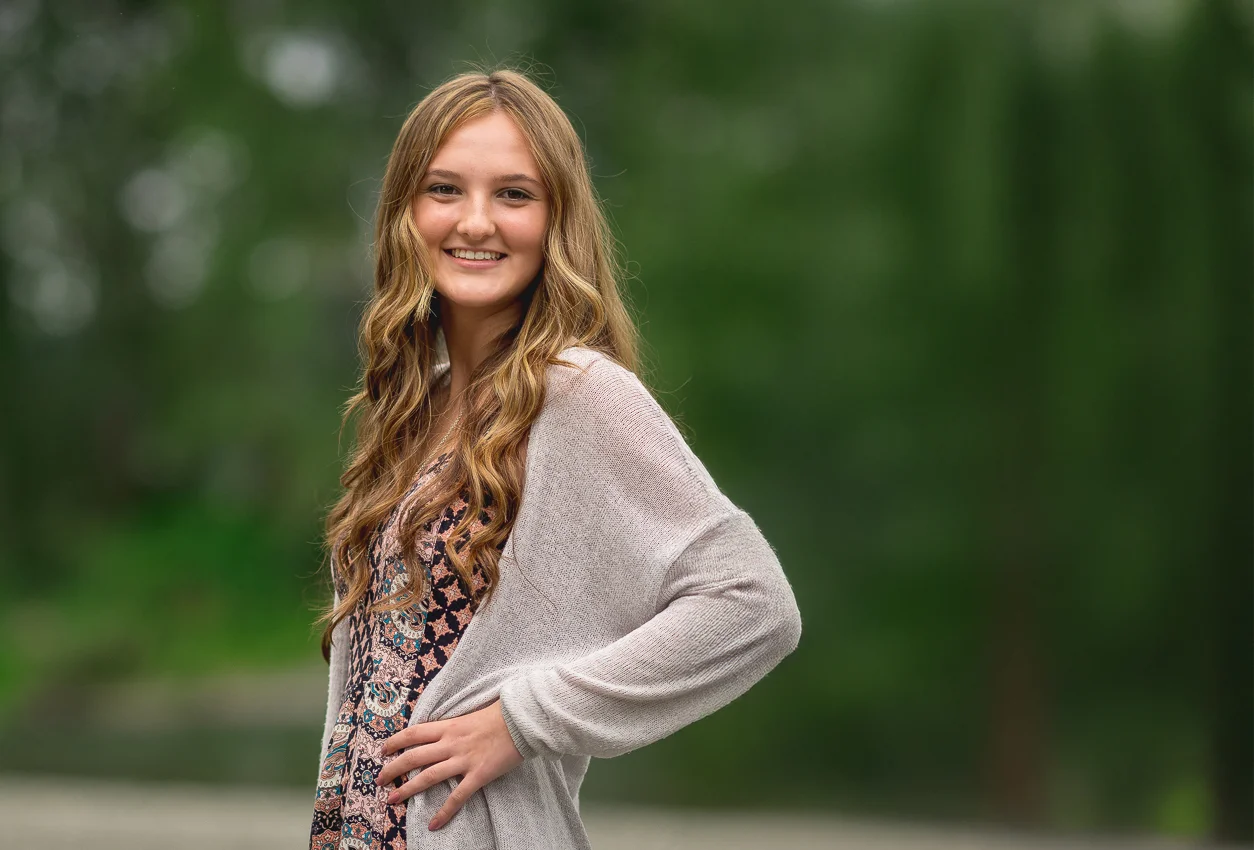 Smiling teen girl with long wavy blonde hair, braces, patterned dress and beige cardigan outdoors