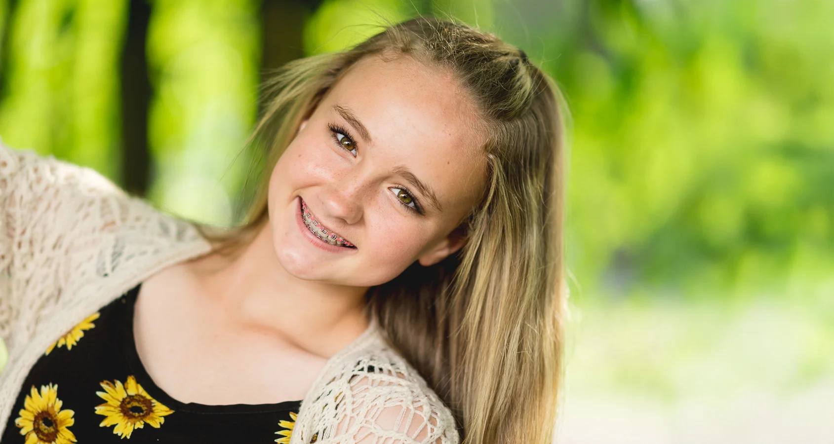 Smiling teen girl with braces, blonde hair, sunflower top, outdoors with green foliage background